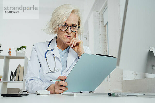 Reifer Arzt untersucht Dokument am Schreibtisch in der Klinik