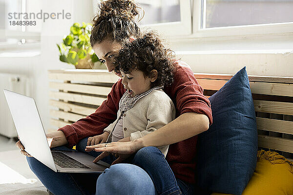 Kleines Mädchen sitzt mit Laptop auf dem Schoß der Mutter auf der Couch zu Hause