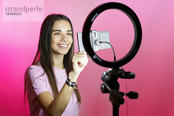 Lächelnder Influencer filmt auf Smartphone im Ringlicht vor rosa Hintergrund
