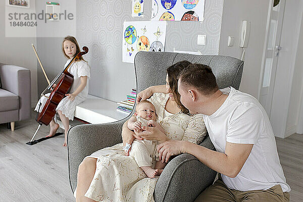 Eltern halten ihren kleinen Jungen und ein Mädchen spielt im Hintergrund ein Musikinstrument