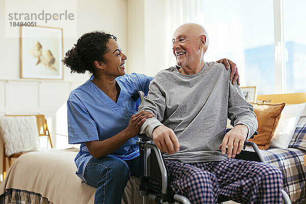Glückliche Krankenschwester mit älterem Mann  der zu Hause im Schlafzimmer im Rollstuhl sitzt