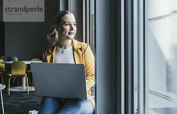 Nachdenkliche Geschäftsfrau mit Laptop schaut aus dem Fenster im Büro