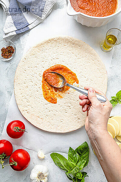Hand of mature woman spreading tomato sauce with spoon on pizza dough