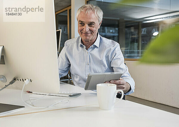 Lächelnder älterer Geschäftsmann mit Tablet-PC und Computer im Büro