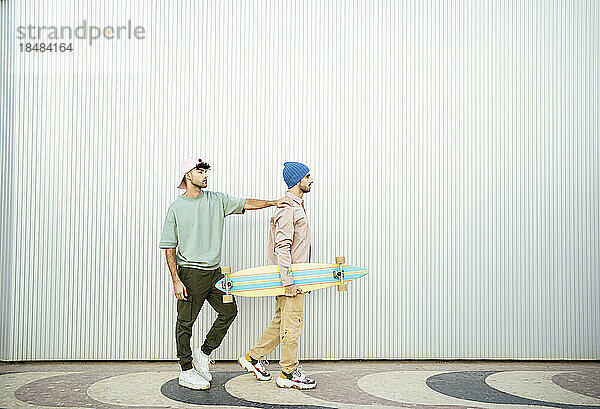 Junger Mann mit Freund hält Longboard vor weißer Wand
