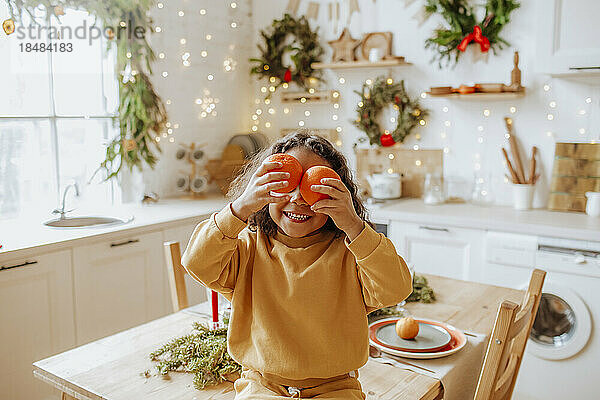 Girl holding oranges over eyes sitting on table at home