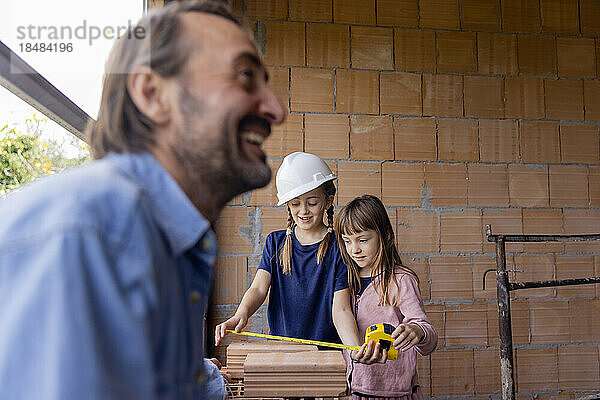 Happy man with daughters measuring bricks in front of wall