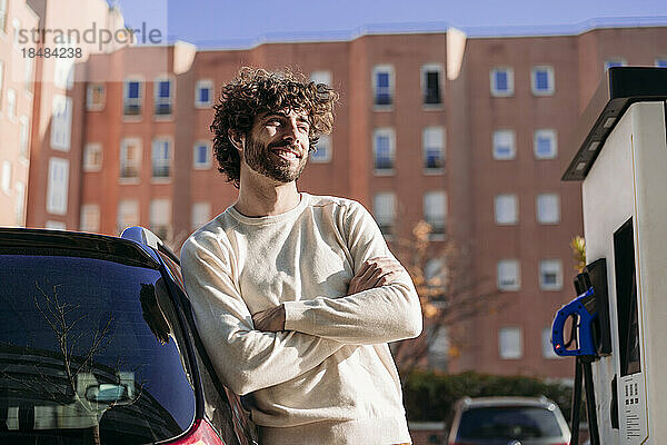 Lächelnder junger Mann stützt sich an der Fahrzeugladestation auf ein Elektroauto