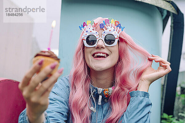 Glückliche Frau mit neuartigen Gläsern  die einen Cupcake in der Hand hält und ihren Geburtstag genießt