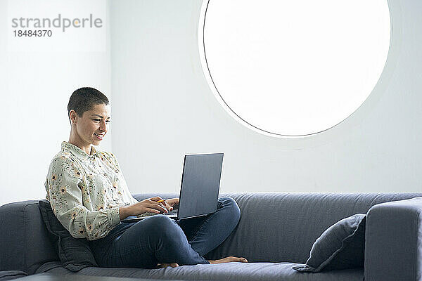 Lächelnde Geschäftsfrau  die am Laptop arbeitet und mit gekreuzten Beinen im Heimbüro sitzt