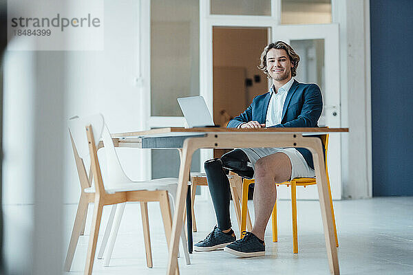 Lächelnder Geschäftsmann sitzt mit Laptop am Schreibtisch im Büro