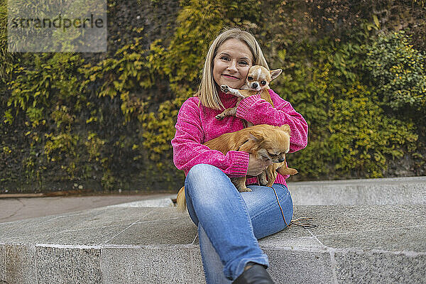 Lächelnde reife Frau sitzt mit Chihuahua-Hunden an der Wand