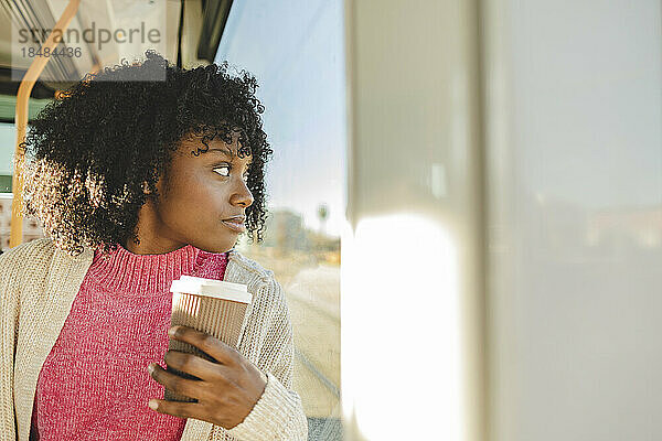 Junge Frau mit Kaffeetasse blickt durch Fenster in Straßenbahn