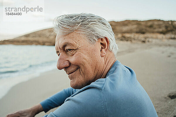 Lächelnder älterer Mann  der bei Sonnenuntergang am Strand sitzt