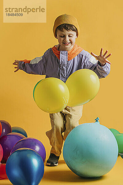Fröhlicher Junge  der mit bunten Luftballons vor gelbem Hintergrund spielt
