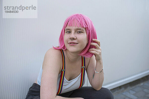 Lächelnde nicht-binäre Person mit rosa Perücke sitzt vor grauer Wand