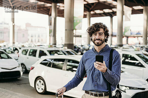 Glücklicher Geschäftsmann mit Smartphone  der vor Autos steht