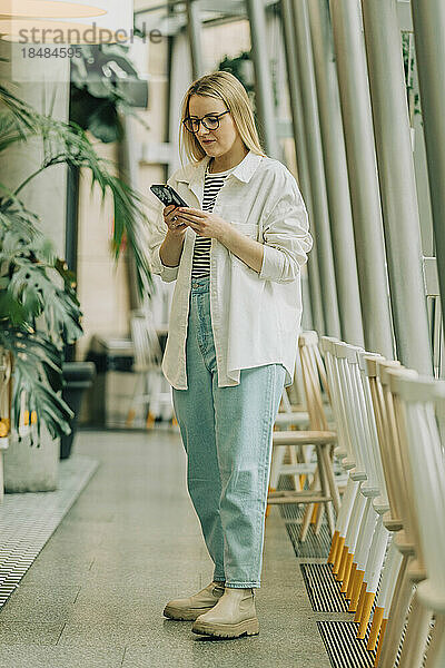 Geschäftsfrau benutzt Smartphone und steht neben Stühlen im Café