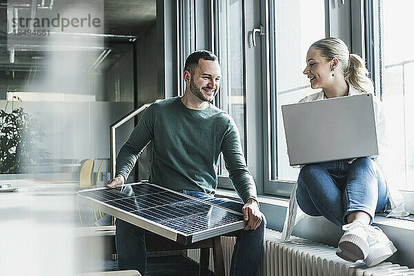 Lächelnder Geschäftsmann hält Solarpanel in der Hand  während sein Kollege im Büro sitzt