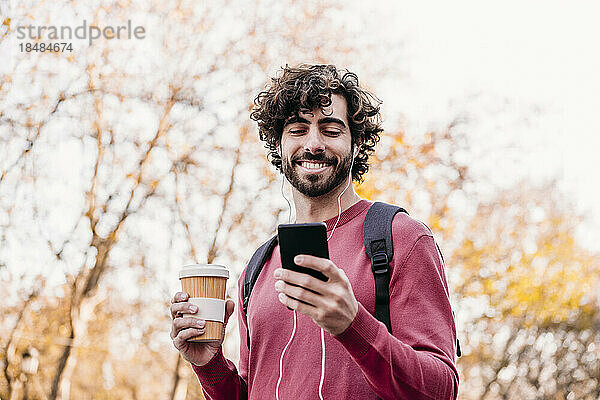 Glücklicher Mann  der sein Mobiltelefon benutzt und eine Kaffeetasse hält  die am Fußweg steht