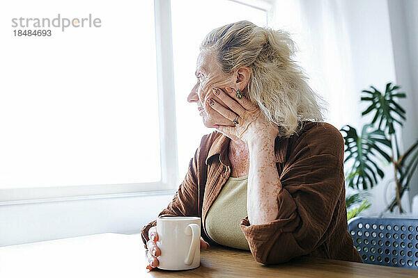 Einsame ältere Frau mit Tasse sitzt zu Hause am Tisch