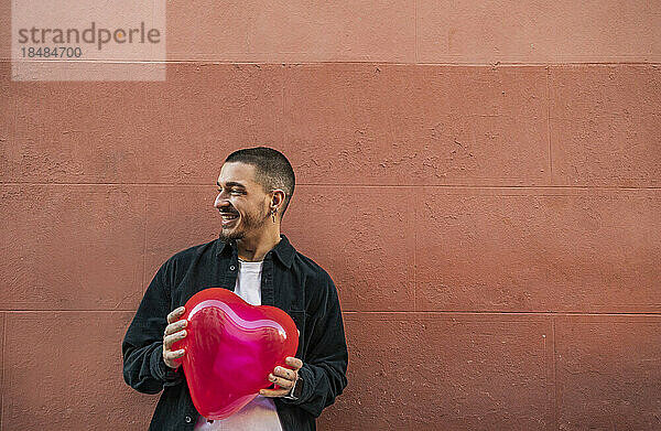 Happy man holding red heart shape balloon in front of wall