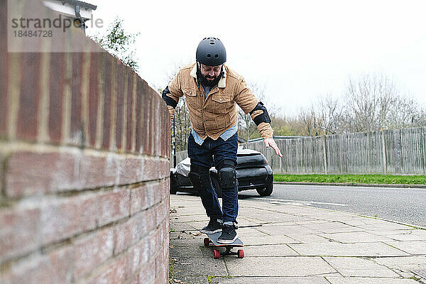 Reifer Mann lernt Skateboardfahren und stützt sich dabei auf die Mauer auf dem Fußweg