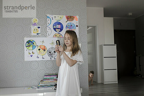 Girl playing with old phone standing by drawings on wall at home
