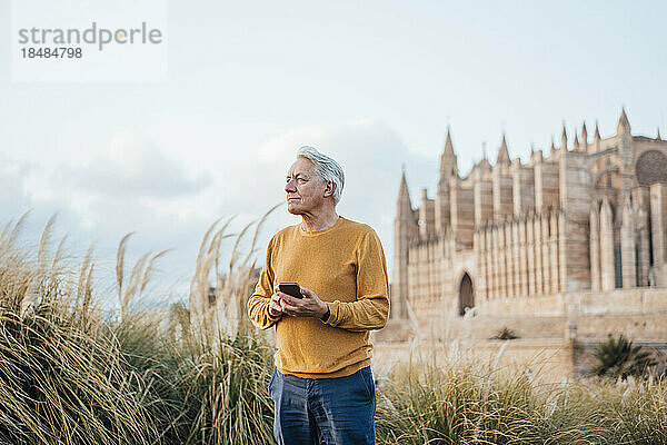 Lächelnder älterer Mann mit Smartphone steht vor dem Gebäude