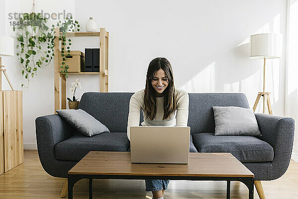 Glückliche junge Frau sitzt mit Laptop auf dem Sofa im Wohnzimmer