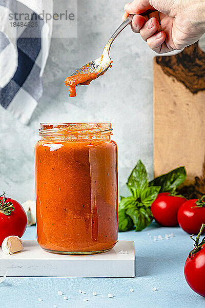 Die Hand einer Frau hält einen Löffel mit Tomatensauce über dem Glas auf dem Tisch