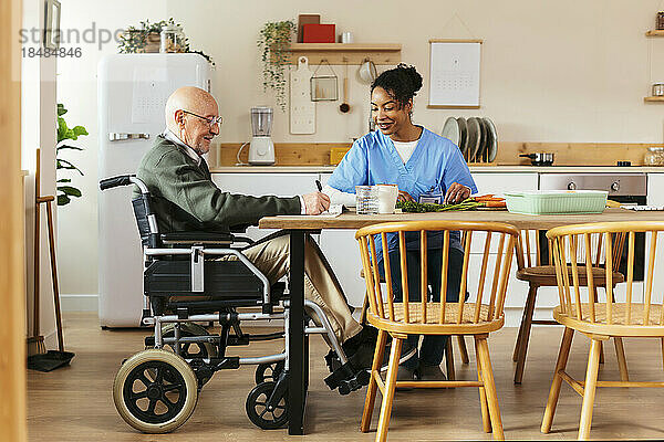 Lächelnder älterer Mann sitzt im Rollstuhl und Hausmeister schneidet zu Hause Gemüse