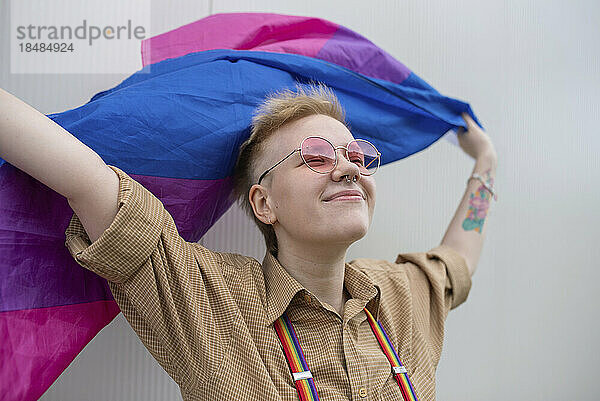 Lächelnde nicht-binäre Person mit Pride-Flagge vor der Wand