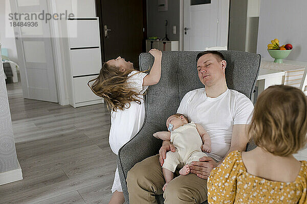 Vater und kleiner Junge entspannen sich auf einem Stuhl  während die Töchter zu Hause spielen
