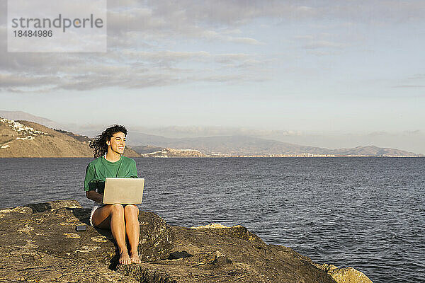 Lächelnde Frau mit Laptop sitzt auf einem Felsen am Meer