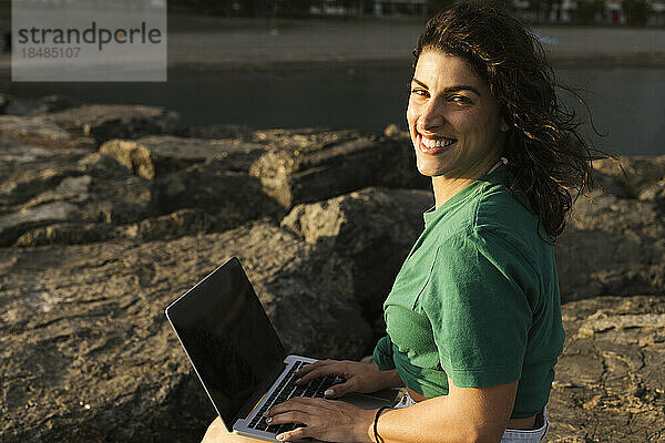 Glückliche Frau mit Laptop sitzt auf einem Felsen am Strand