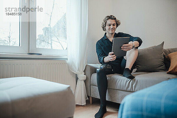 Junger Mann sitzt zu Hause auf dem Sofa und benutzt einen Tablet-PC