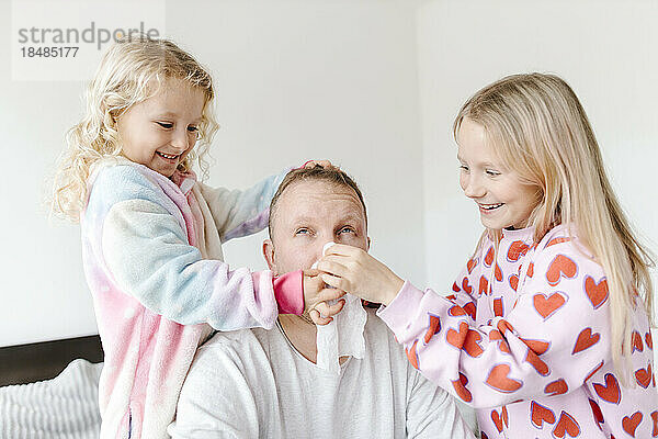 Glückliche Töchter wischen ihrem Vater zu Hause mit einer Serviette die Nase ab
