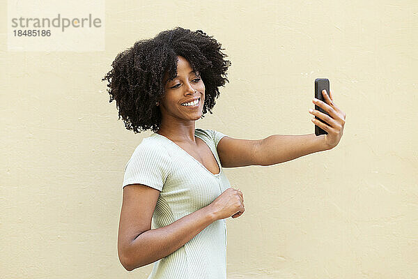 Lächelnde junge Frau macht ein Selfie mit ihrem Smartphone an einer beigen Wand