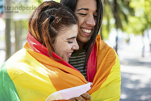 Happy lesbian couple covered in rainbow colored flag on footpath