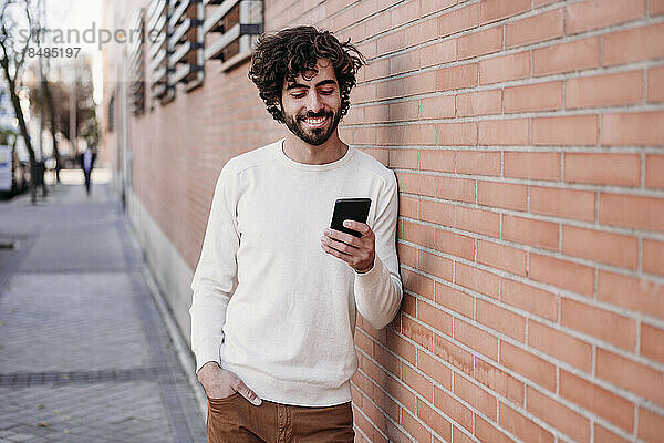 Glücklicher junger Mann benutzt Smartphone und lehnt an der Wand