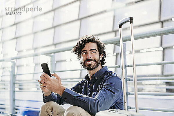 Lächelnder junger Geschäftsmann sitzt mit Smartphone am Bahnhof