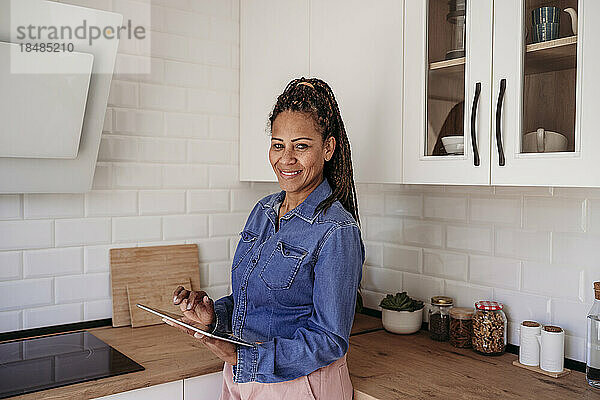 Glückliche reife Frau mit Tablet-PC  die zu Hause in der Küche steht