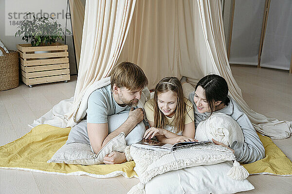 Lächelnde Eltern und Tochter lesen Buch im Deckenzelt