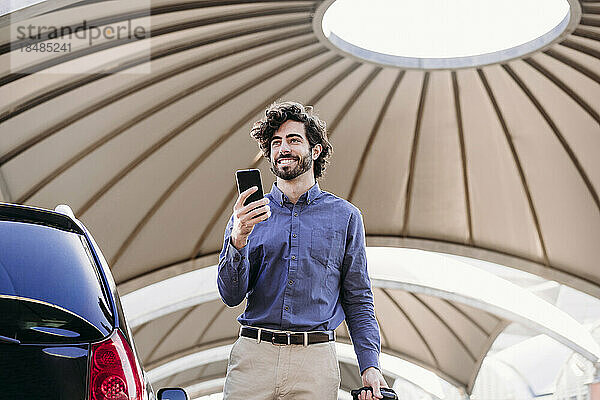 Glücklicher junger Geschäftsmann mit Smartphone  der auf dem Parkplatz steht
