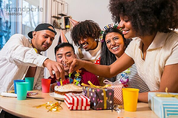 Multiethnische Gruppe von Freunden bei einer Geburtstagsfeier auf dem Sofa zu Hause mit einem Kuchen und Geschenken  lächelnd Platzierung der Happy Britday Kerzen