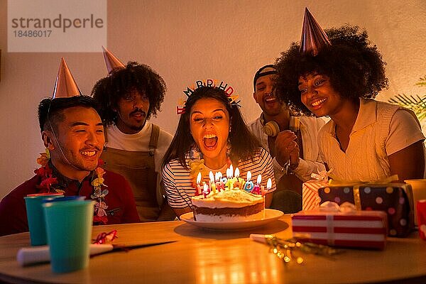 Multiethnische Gruppe von Freunden bei einer Geburtstagsfeier auf dem heimischen Sofa mit Kuchen und Geschenken  Ausblasen der Kerzen bei ausgeschaltetem Licht