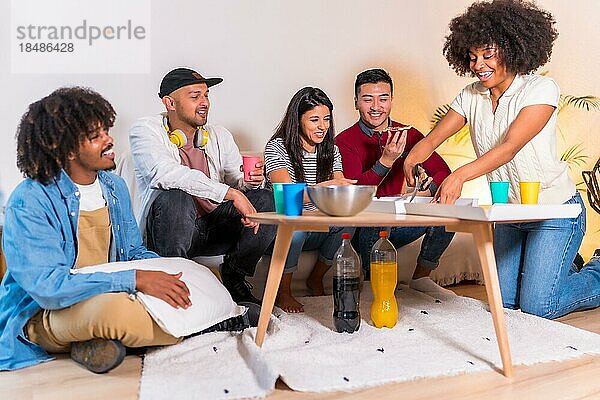 Gruppe multiethnischer Freunde auf einem Sofa  die auf einer Party zu Hause Pizza essen und alkoholfreie Getränke trinken  die Pizza aufschneiden und sie mit Freunden teilen