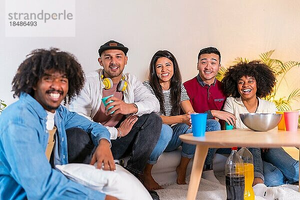 Gruppenporträt multiethnischer Freunde  die auf dem Sofa Popcorn essen und Limonade trinken und lächelnd in die Kamera schauen
