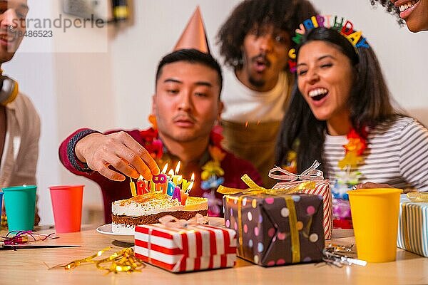 Multiethnische Gruppe von Freunden bei einer Geburtstagsfeier auf dem Sofa zu Hause mit einem Kuchen und Geschenken  chinesischer Mann zündet die Kerzen an